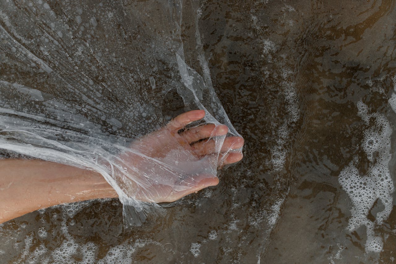 creative-03 A hand holds a plastic sheet amidst seawater foam on a sandy beach, highlighting pollution.